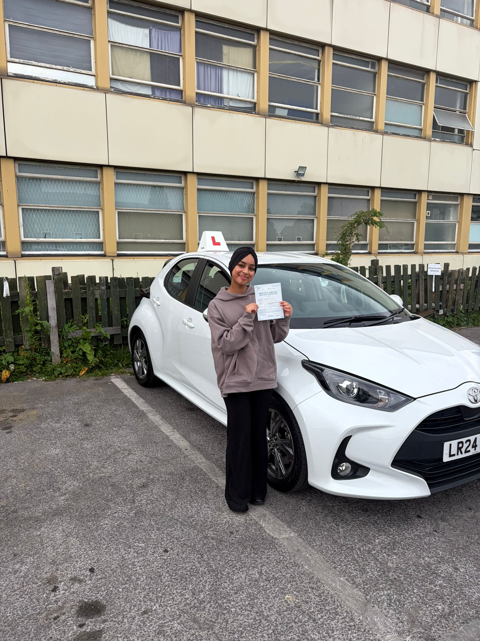 Passed student holding a driving test pass certificate beside a white learner car