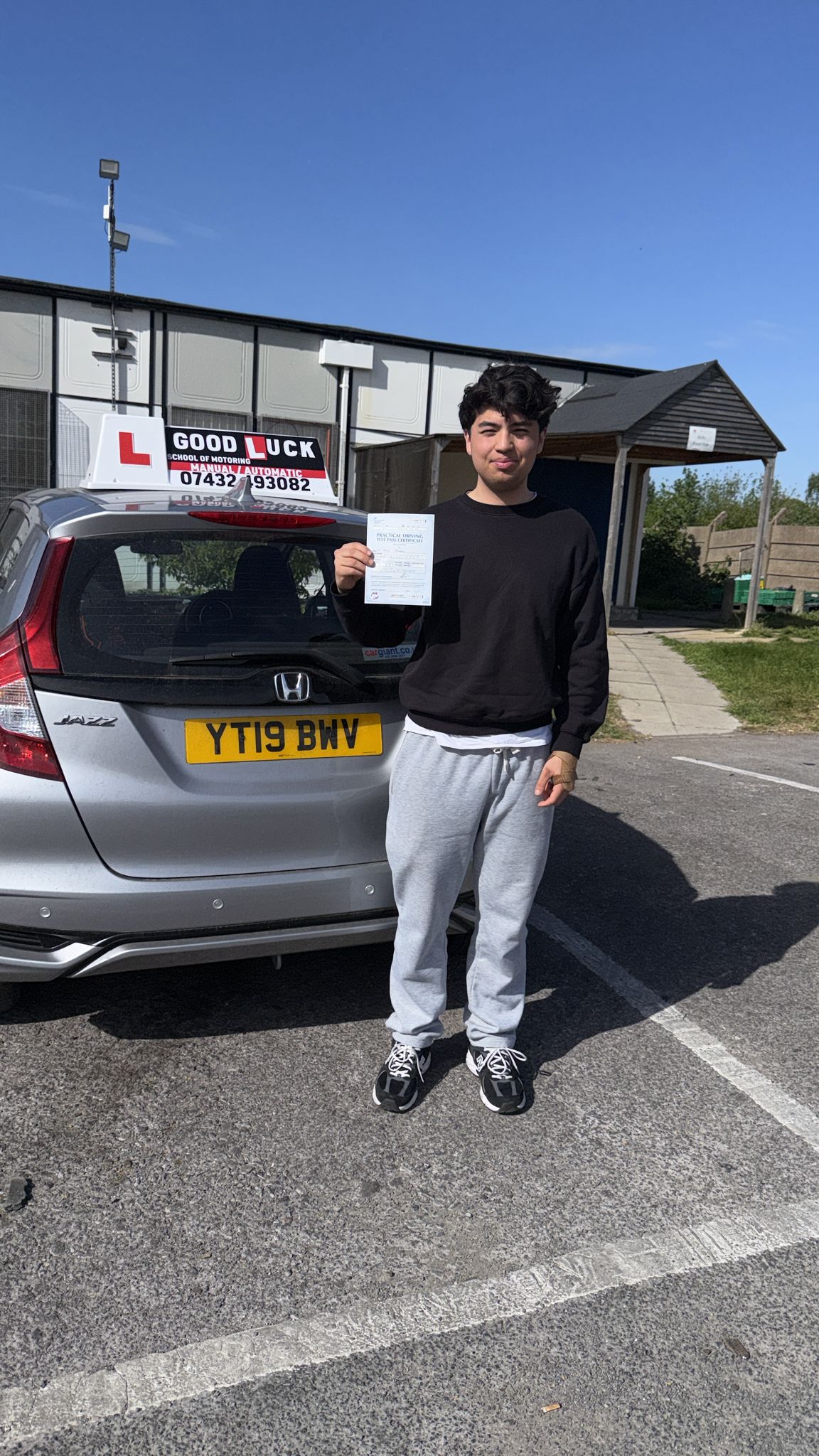 Passed student holding a driving test pass certificate beside a Good Luck learner car