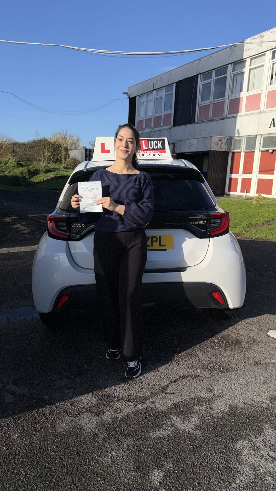 Passed student holding a driving test pass certificate beside a Good Luck learner car